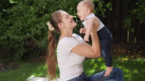 Feliz madre alegre sosteniendo y levantando a su pequeño hijo en el jardín del patio trasero. Crianza, familia, desarrollo infantil y diversión al aire libre en la naturaleza. — Vídeos de Stock