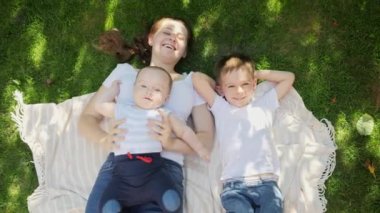 Mutlu çocuklar ve anneleri parkta çimlerde dinlenip yukarı bakıyorlar. Ebeveynlik, aile, çocuk gelişimi ve doğada eğlence..