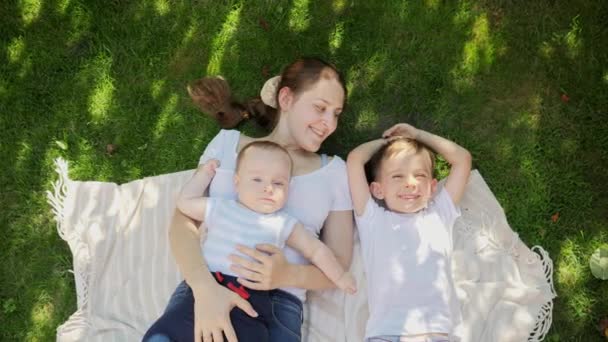Vue de dessus de la mère souriante avec deux fils couchés sur la couverture sur l'herbe et regardant dans le ciel. Parentalité, famille, développement des enfants et plaisir en plein air dans la nature. — Video