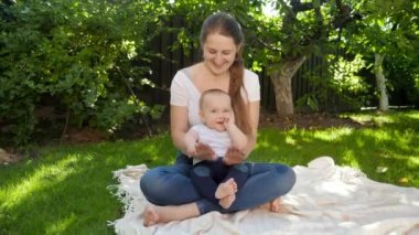 Gülümseyen bir annenin portresi, çimde oturan ve kameraya gülümseyen bir bebekle birlikte. Ebeveynlik, aile, çocuk gelişimi ve doğada eğlence..
