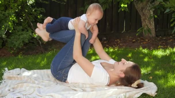 Gelukkige glimlachende moeder die haar benen vasthoudt en haar zoontje optilt in de tuin. Ouderschap, familie, ontwikkeling van kinderen en plezier in de natuur. — Stockvideo