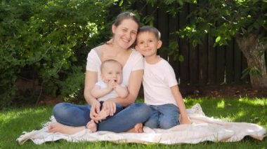 Parkta çimlerin üzerinde oturan ve kameraya gülümseyen iki oğlu olan mutlu bir anne. Ebeveynlik, aile, çocuk gelişimi ve doğada eğlence..