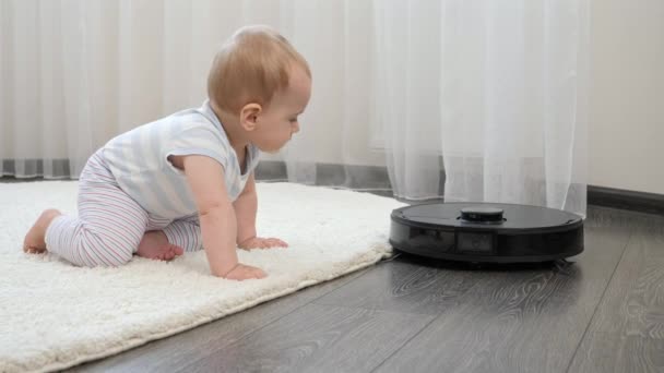 Schattige baby jongen op zoek met interesse voor robot stofzuiger op de vloer in de woonkamer. Concept van hygiëne, huishoudelijke apparaten en robots in het moderne leven. — Stockvideo