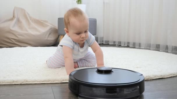 かわいい赤ちゃんの男の子が床に這い、ロボット掃除機を見ています。現代生活における衛生、家庭用品、ロボットの概念. — ストック動画