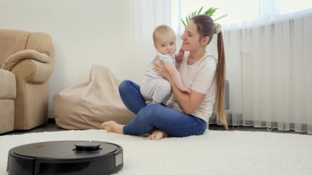 Šťastná usměvavá matka si hraje se svým malým synem, zatímco robot vysavač čistý dům. Koncepce hygieny, domácích pomůcek a robotů v moderním životě. — Stock video