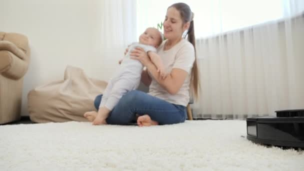 Staubsaugerroboter arbeiten neben Baby, das mit Mutter auf Teppich im Haus spielt. Hygienekonzept, Haushaltsgeräte und Roboter im modernen Leben. — Stockvideo