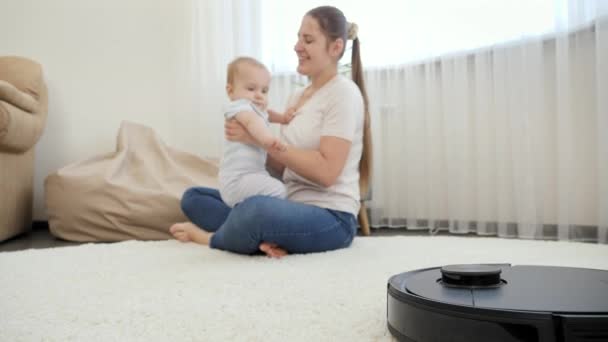 Aspirador de robô ajudando a mulher fazendo tarefas enquanto ela passa o tempo com o bebê filho. Conceito de higiene, aparelhos domésticos e robôs na vida moderna. — Vídeo de Stock