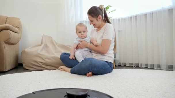 Szczęśliwa uśmiechnięta matka z synkiem patrzącym na odkurzacz robota czyszczący dywan i podłogę w domu. Koncepcja higieny, gadżetów gospodarstwa domowego i robotów we współczesnym życiu. — Wideo stockowe