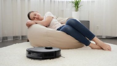 Oturma odasında çalışan robot elektrik süpürgesinin yanında uyuyan genç kadın. Modern hayatta hijyen, ev aletleri ve robotlar kavramı.