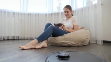 Robot elektrik süpürgesi ev işi yaparken genç bir kadın puf koltukta akıllı telefonuyla dinleniyor. Modern hayatta hijyen, ev aletleri ve robotlar kavramı.