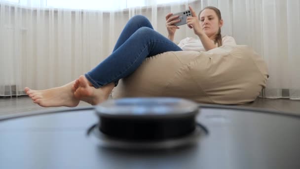 로봇 청소기 의 POV 샷 집 청소를 하는 동안젊은 여성은 휴식을 취하고 스마트폰을 사용 한다. 현대 생활 에서의 위생, 가정용품 및 로봇 의 개념. — 비디오
