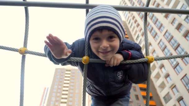 Porträtt av söta leende pojke klättra upp i rep nätet och vinka i kameran. Begreppet barnutveckling, idrott och utbildning. — Stockvideo
