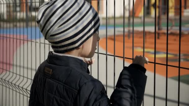 Upset stressed little boy holding and looking through high metal fence on expensive school and playground. Concept of poverty, immigration, bullying and kids stress — Stock Video