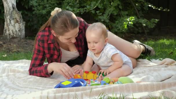 Carino bambino sorridente con la giovane madre che gioca giocattoli sull'erba nel parco. Concetto di sviluppo del bambino, famiglia che ha tempo insieme e genitorialità. — Video Stock