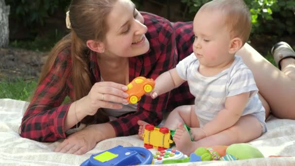 Gelukkige lachende moeder met kleine baby zoon die speelgoed speelt op gras in het park. Concept van de ontwikkeling van het kind, familie die samen tijd doorbrengt en ouderschap. — Stockvideo