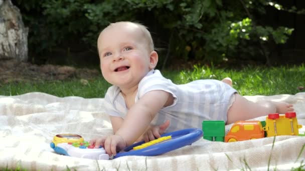 Schattig lachend jongetje liggend op gras in de tuin en spelend met kleurrijk speelgoed. Begrip "vroegtijdige ontwikkeling van het kind", "onderwijs" en "ontspannen buitenshuis". — Stockvideo