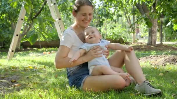 リンゴの木の果樹園や庭で草の上に座っている母親と幸せな笑顔の赤ちゃん。子供の早期開発、教育、屋外でのリラックスの概念. — ストック動画