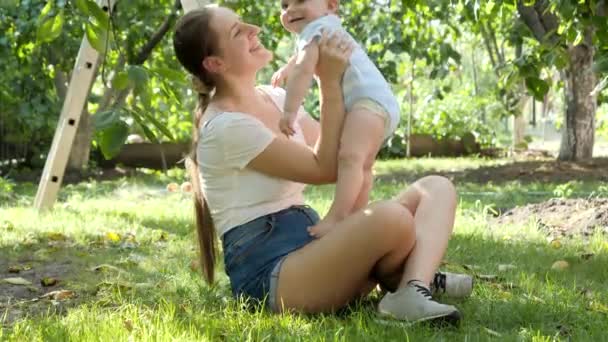 Boldog mosolygó anya felemeli és megcsókolja a kisfiát az almafák alatt a gyümölcsösben. A gyermekek korai fejlődésének, oktatásának és kikapcsolódásának koncepciója a szabadban. — Stock videók