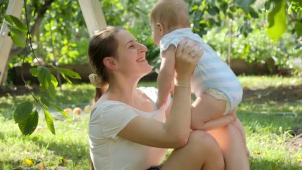 Portret van een lachende liefhebbende moeder die haar zoontje knuffelt terwijl ze zich ontspant op gras in de tuin. Concept van de ontwikkeling van het kind, familie die samen tijd doorbrengt en ouderschap. — Stockvideo