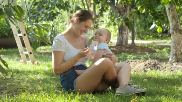 Glimlachende moeder geeft boomblad aan haar kleine zoontje terwijl ze op gras zit onder appelboom in boomgaard. Concept van de ontwikkeling van het kind, familie die samen tijd doorbrengt en ouderschap. — Stockvideo