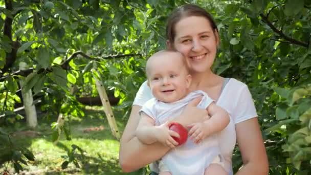 Πορτρέτο του χαμογελαστού μωρού με τη μητέρα κρατώντας ώριμα μήλα και ποζάροντας στον κήπο πίσω αυλή. Έννοια της ανάπτυξης παιδιών, της γονικής μέριμνας και της υγιούς βιολογικής καλλιέργειας τροφίμων. — Αρχείο Βίντεο