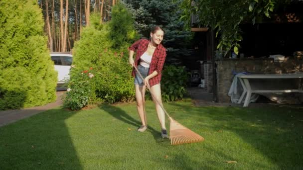Eine junge lächelnde Frau arbeitet im Hinterhof des Hauses und sammelt mit der Gartenharke umgefallenes Laub ein. Konzept der Hausarbeit, Gartenarbeit und des Vorortlebens — Stockvideo