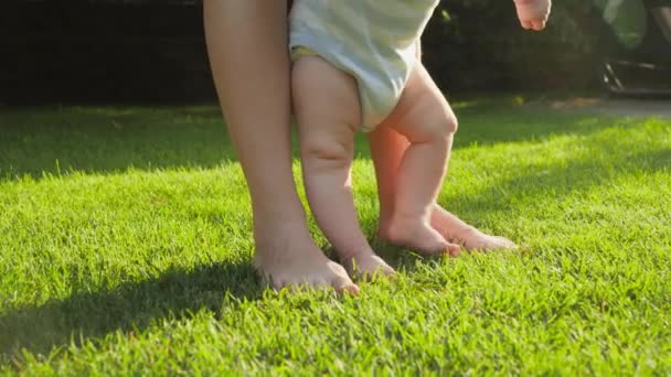 家の裏庭の庭で新鮮な緑の芝生の芝生の上に立っている母親と裸足の赤ちゃんの閉鎖。健康的なライフスタイル、子供の発達と子育ての概念. — ストック動画