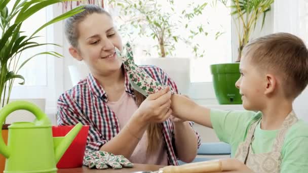 Ung mamma hjälper sin son att sätta trädgårdshandskar innan hon planterar blommor i krukor hemma. Begreppet trädgårdsskötsel, hemplantering och familjearbete tillsammans. — Stockvideo