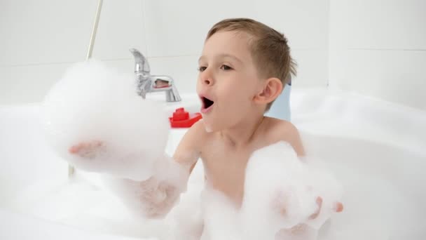 Lächelnder kleiner Junge, der mit Seifenschaum in der Badewanne spielt und ihn von den Händen bläst. Konzept der Hygiene, Entwicklung der Kinder und Spaß zu Hause — Stockvideo