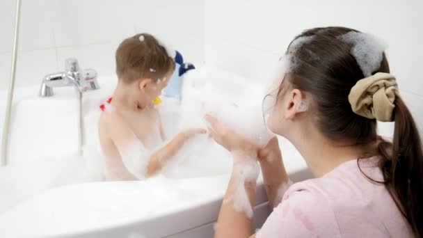 Mãe engraçada com o filho espirrando água no banho e jogando sabão suds. Conceito de hygine, desenvolvimento infantil e diversão em casa — Vídeo de Stock