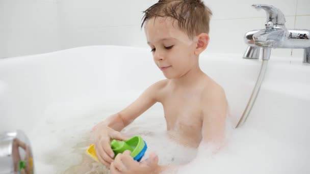 Χαριτωμένο αγόρι κάθεται στο μπάνιο με αφρό σαπουνιού και παίζει παιχνίδια βάρκες και πλοία. Έννοια του οικογενειακού χρόνου, ανάπτυξη παιδιών και διασκέδαση στο σπίτι — Αρχείο Βίντεο