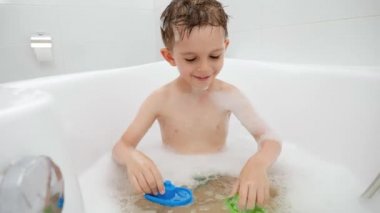 Oyuncak teknelerle oynayan ve banyoda su sıçratan neşeli güler yüzlü çocuk. Aile zamanı kavramı, çocuk gelişimi ve evde eğlence
