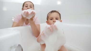 Annesinin oynadığı ve sabun köpüğüyle eğlendiği pozitif gülümseyen bir çocuk. Aile zamanı kavramı, çocuk gelişimi ve evde eğlence
