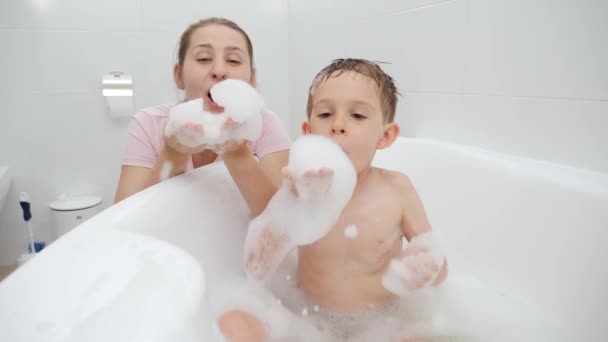 Cheerful laughing mother and little son blowing soap bubbles and foam while playing in bathroom. Concept of family time, children development and fun at home — Stock Video