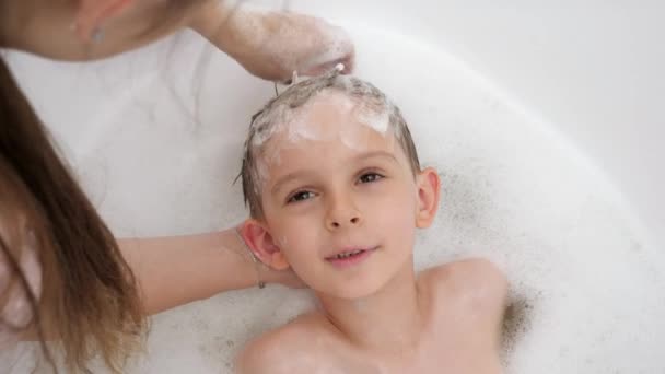 Портрет усміхненого маленького хлопчика, який дивиться на матір, коли вона миє волосся шампунем у ванній. Концепція гігієни, розвиток дітей та розваги вдома — стокове відео