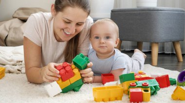 Gülümseyen anne, bebeğiyle birlikte halıda yatıyor ve onunla oyuncaklar oynuyor.