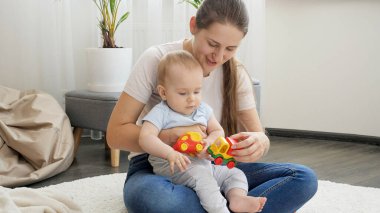 Genç, şefkatli bir anne, küçük oğluyla oturma odasında halının üzerinde oyuncak oynuyor.