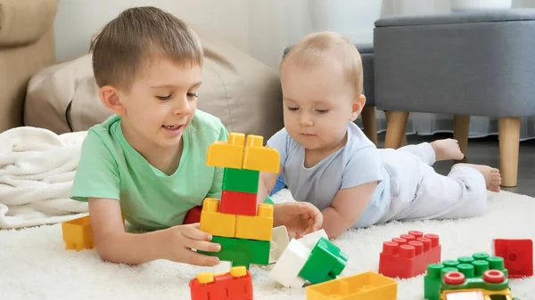 Chlapec učí svého bratříčka stavět věž s kostkami a cihlami. Koncepce vývoje, vzdělávání a tvořivosti dětí doma — Stock fotografie