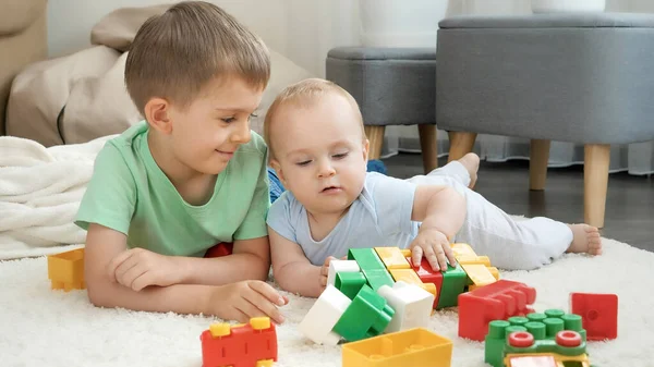 Uśmiechnięty chłopiec patrząc na swojego braciszka bawiącego się klockami i klockami na podłodze w pokoju zabaw — Zdjęcie stockowe