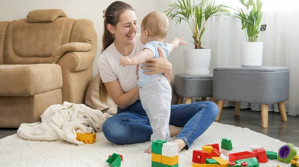 Feliz madre sonriente apoyando a su pequeño hijo bebé y enseñando a caminar en la alfombra en la sala de estar. Concepto de desarrollo infantil, educación y creatividad en el hogar — Foto de Stock