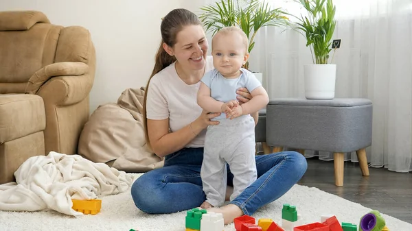 Portrait of happy smiling mother supporting her little baby son standing on acrpet in living room — Stock Photo, Image