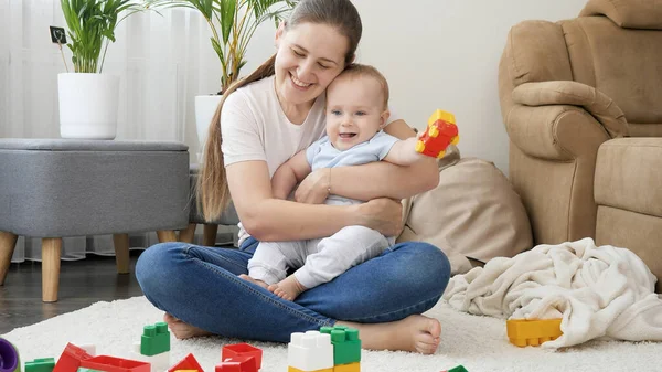Молодая улыбающаяся мать обнимает и держит своего маленького сына, играя с игрушечными машинками на полу в гостиной. Концепция семейного времяпровождения и развития детей — стоковое фото