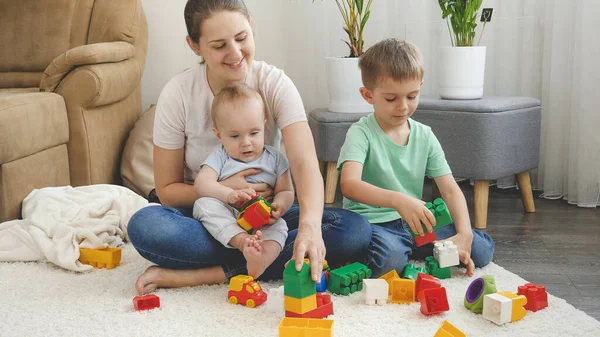 Pequeño niño y hermano mayor jugando con la madre en juguetes en la alfombra en la sala de estar. Concepto de familia teniendo tiempo juntos y desarrollo de los niños — Foto de Stock