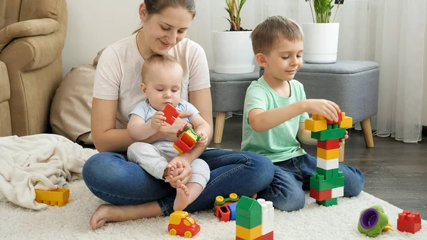 カーペットで遊んでおもちゃの塔を構築する母親と幸せな子供たち。家族の時間と子供の発達の概念 — ストック写真
