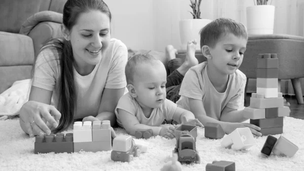 Czarno-biały portret szczęśliwych dzieci z matką leżącą na dywanie w salonie i bawiącą się zabawkami. — Zdjęcie stockowe