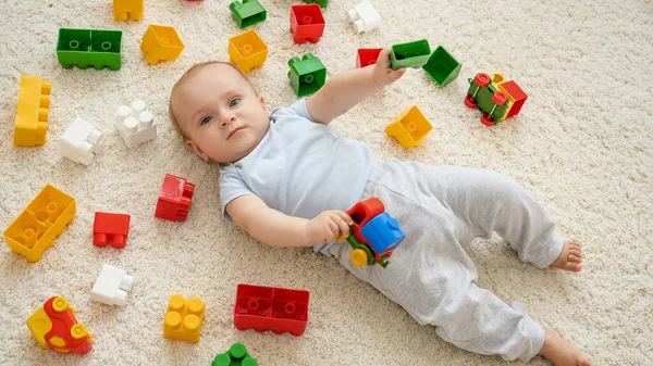 Malý usměvavý chlapeček na koberci v herně pokrytý barevnými hračkami, cihlami a bloky. Koncepce vývoje, vzdělávání a tvořivosti dětí doma — Stock fotografie