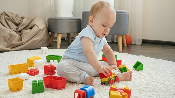 Aranyos kisfiú ül a szőnyegen, és játszik színes műanyag játékautóval. A gyermekek fejlődésének, oktatásának és kreativitásának fogalma otthon — Stock Fotó