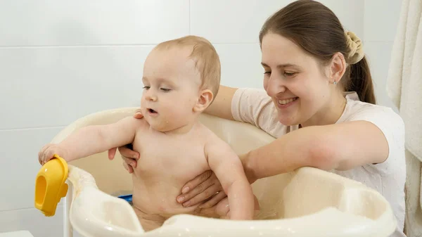 Feliz madre sonriente mirando a su pequeño hijo salpicando agua y jugando en el baño. Concepto de higiene infantil, atención sanitaria y crianza de los hijos. — Foto de Stock