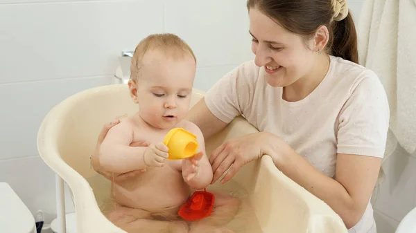 Щаслива усміхнена мати дивиться, як її дитина миє у ванній кімнаті і грає в іграшки — стокове фото