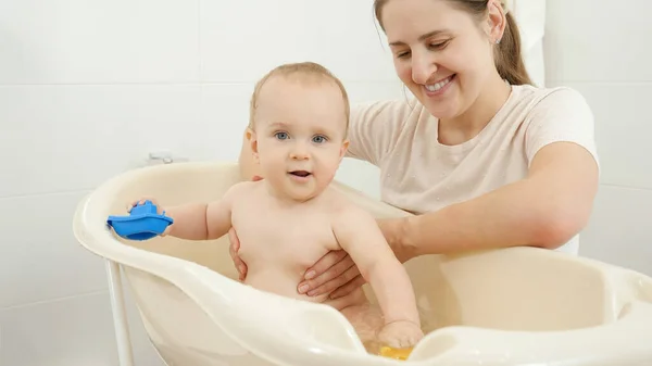 Madre sonriente sosteniendo y mirando a su hijo pequeño jugando en el baño de niños pequeños en casa. Concepto de higiene infantil, atención sanitaria y crianza de los hijos. — Foto de Stock